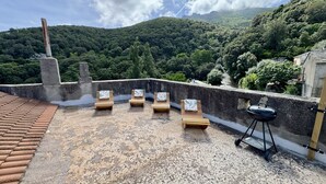A rooftop terrace with wooden lounge chairs, a barbecue grill, and a view of a lush green hillside.