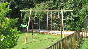 A playground with swings and a slide, surrounded by a wooden fence and greenery.