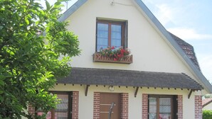 A two-story house with a sloped roof, a front door, and a window with shutters. There is a barbecue grill and outdoor furniture visible.