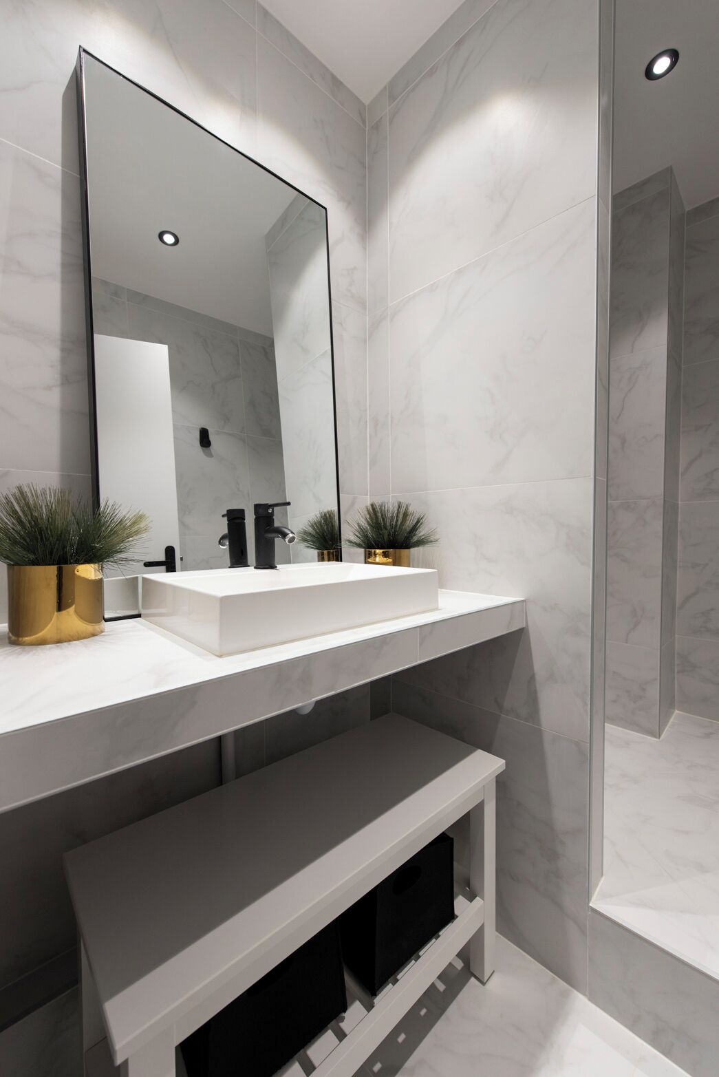 A modern bathroom with a white vanity, two gold potted plants, a large mirror, and a shower area.