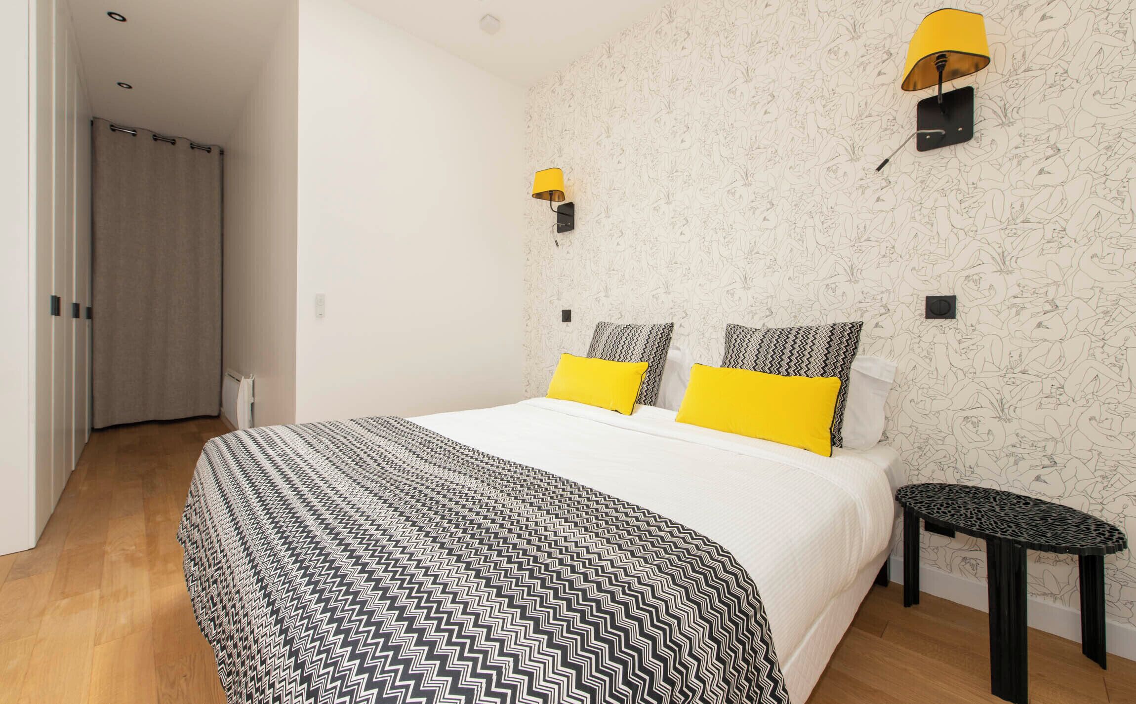 Une chambre à coucher avec une couvre-lit à motifs, deux oreillers, une petite table de chevet noire et une lampe fixée au mur.