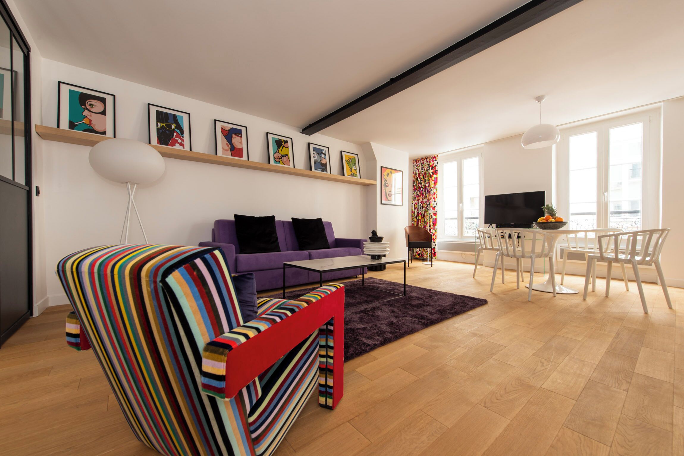 A modern living room with a striped sofa, a purple couch, a flat-screen TV, and a dining area with white chairs.