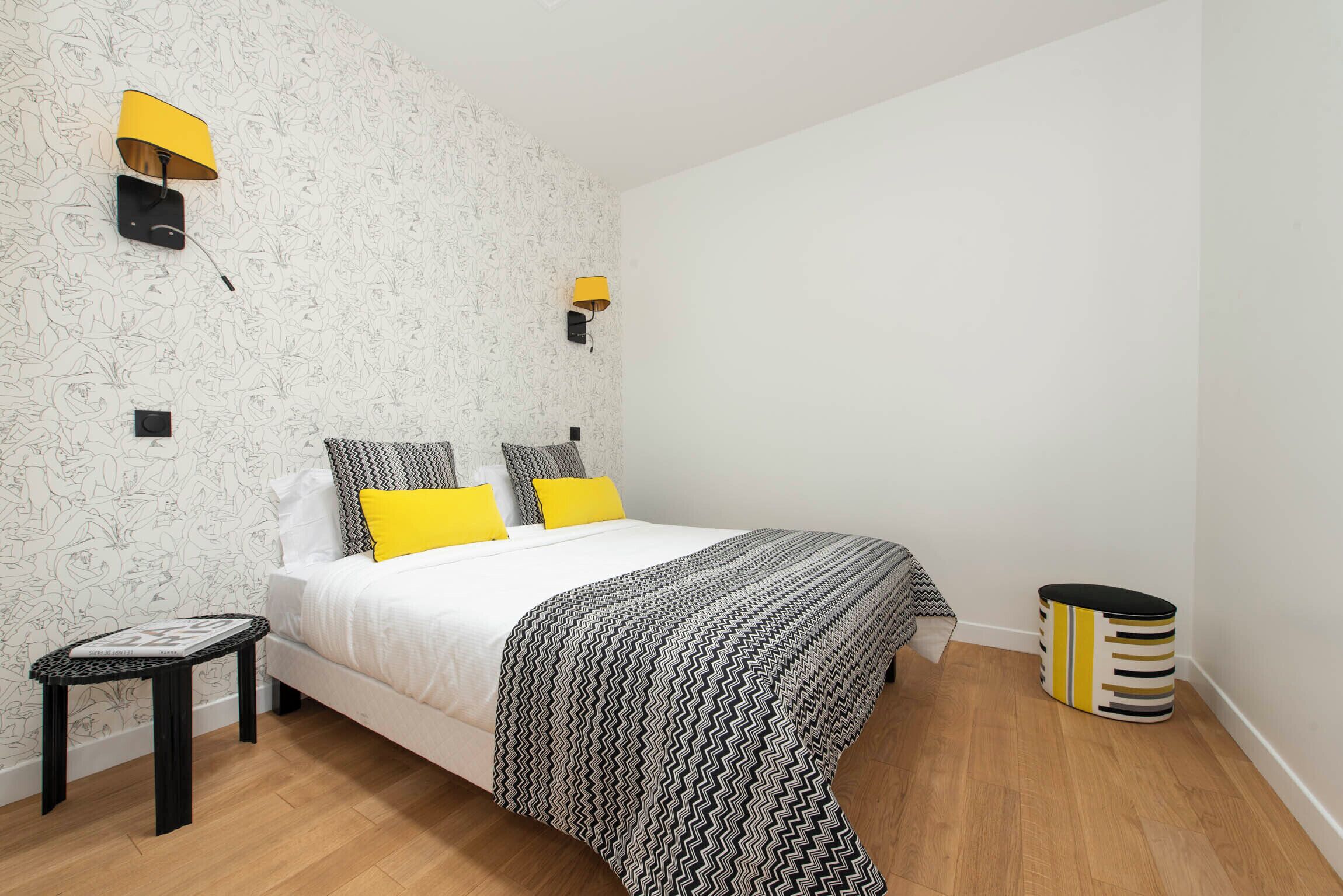 Un dormitorio con papel tapiz estampado, una cama con ropa de cama blanca y amarilla, dos lámparas fijadas a la pared, una mesita de noche pequeña y un cubo de basura cilíndrico.