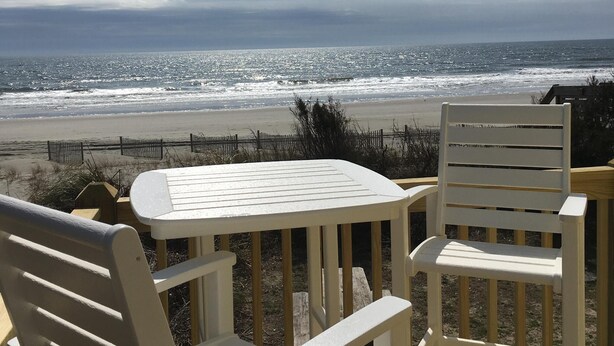 Une terrasse avec des chaises blanches et une table, donnant sur une plage où l’on peut voir des vagues et un ciel nuageux.