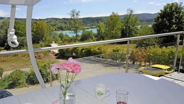 Un balcon avec une table dressée pour deux personnes, un vase rempli de fleurs roses et une vue sur un lac et une forêt.