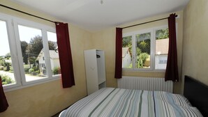 Une chambre à coucher avec un lit, deux fenêtres avec des rideaux, un radiateur et une vue sur une maison et de la verdure à l’extérieur.
