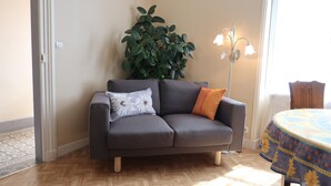 Ein graues Sofa mit zwei Kissen, einer Topfpflanze und einem Tisch mit gemustertem Tischtuch.