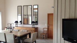 A modern living room with a dining area, a flat-screen TV mounted on a wooden wall, and a large mirror above the dining table. - House with calm 140m ² with swimming pool 10x5 (Campagne)