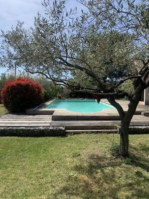 Une piscine avec une terrasse en bois, entourée de pelouse et d’un arbre.