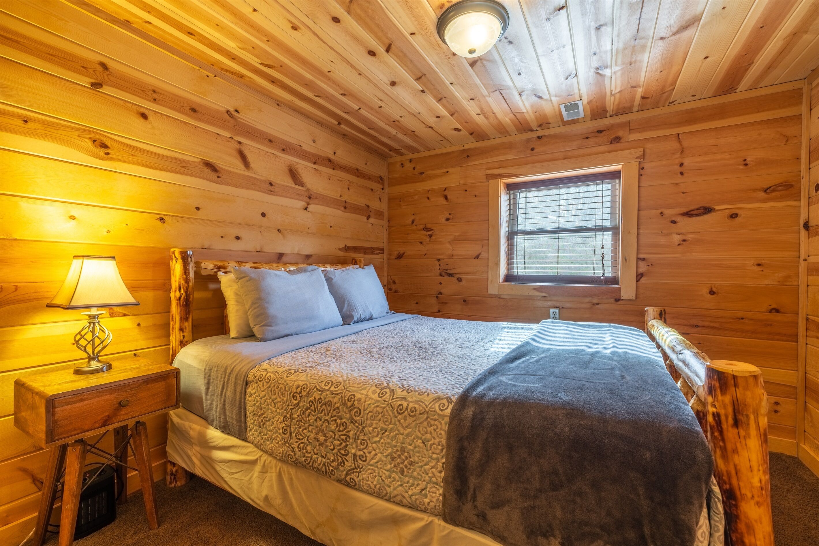 A wooden cabin bedroom with a bed, a nightstand, a lamp, and a window.