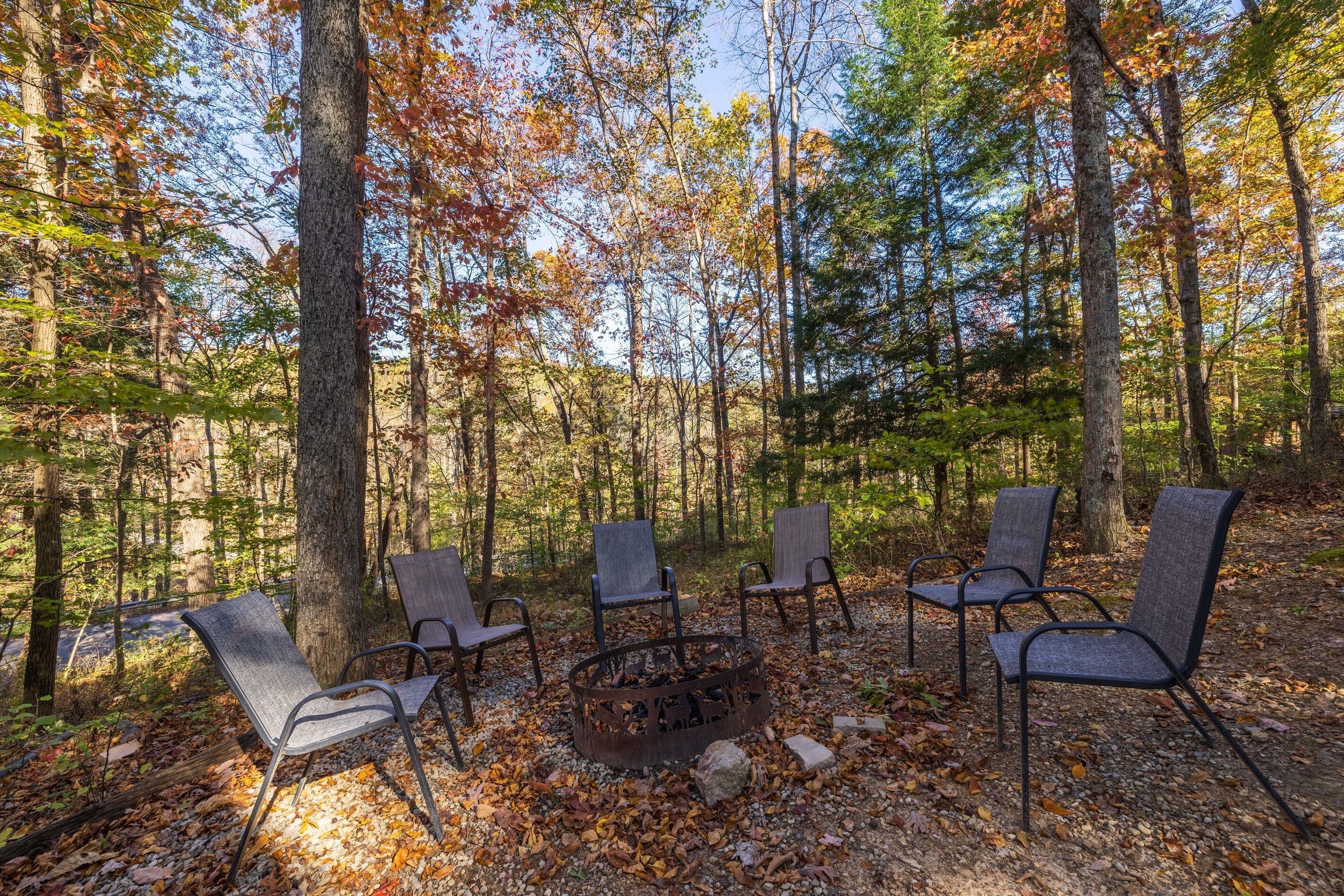 A forest setting with a fire pit and five chairs arranged around it.