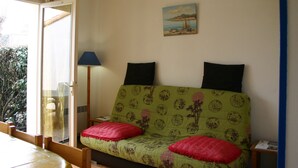 A room with a green sofa with red cushions, a painting of a bridge, and a window with a view of trees. - Cozy Vacation Home Near Beach with Garden, Parking, and WiFi (La Tranche-sur-Mer)