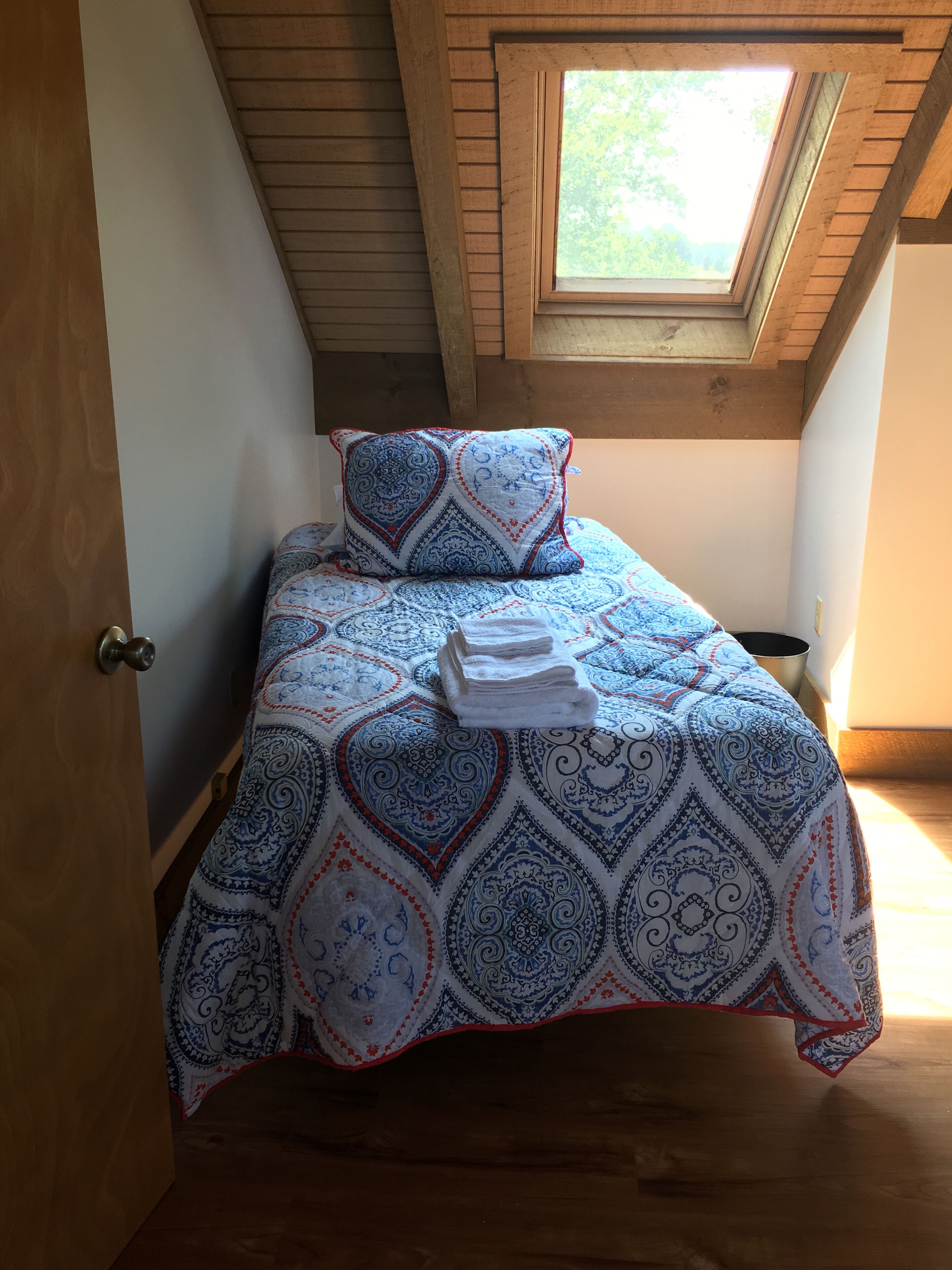A small, cozy bedroom with a sloped ceiling and a skylight, featuring a bed with a patterned bedspread and matching pillow, and a neatly folded towel on the bed.