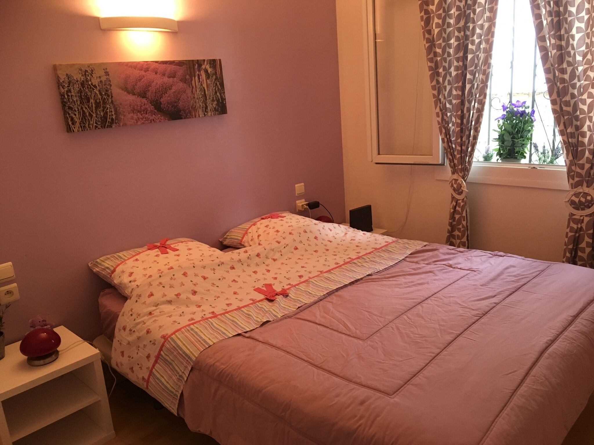 A bedroom with a bed, a nightstand, a wall-mounted lamp, and a window with curtains.