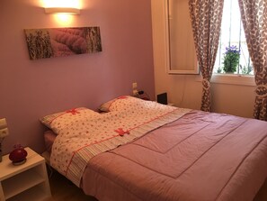 Un dormitorio con cama, mesita de noche, lámpara de pared y ventana con cortinas.