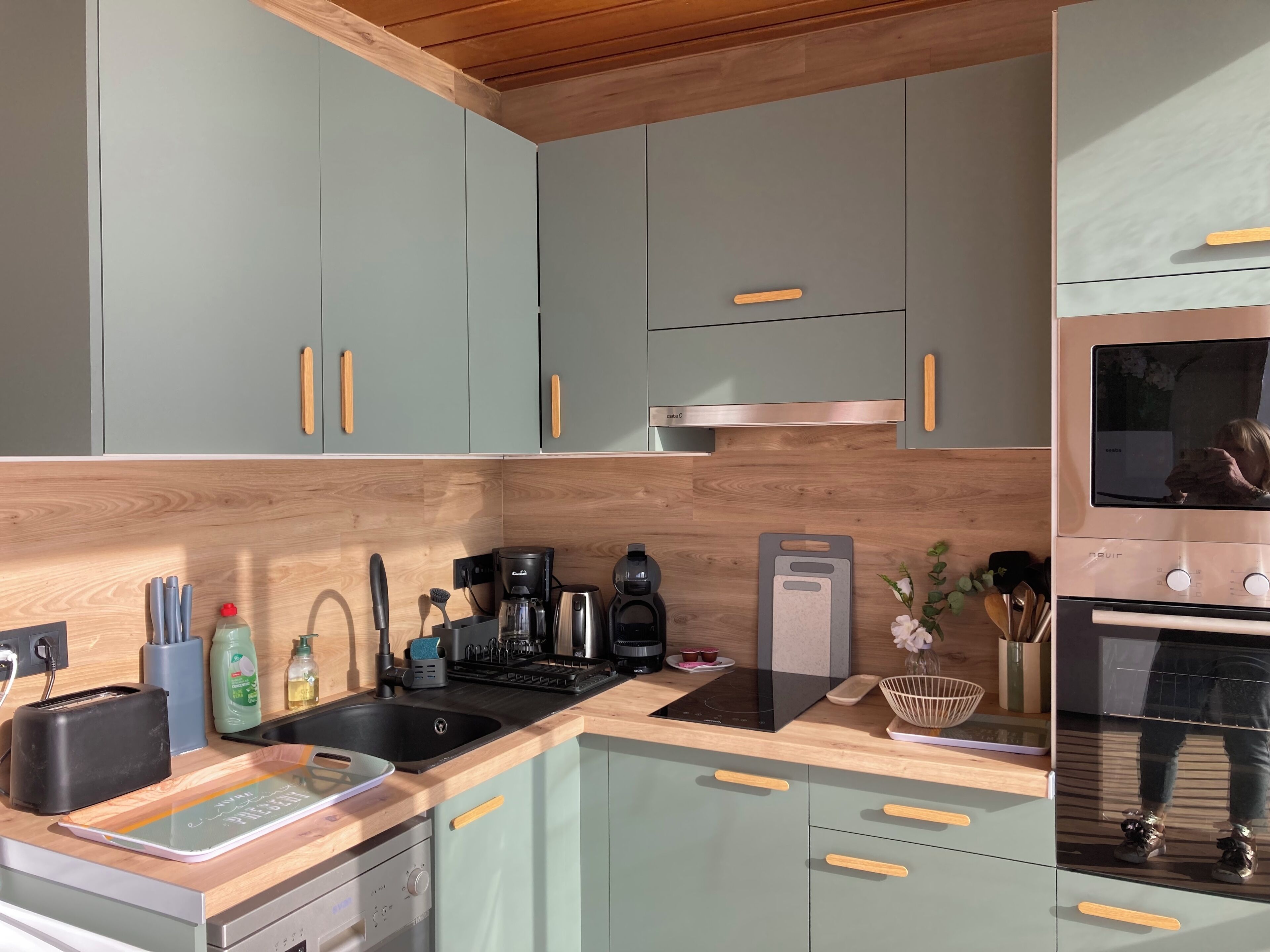 A modern kitchen with light green cabinets, wooden handles, a stainless steel sink, and a built-in oven.
