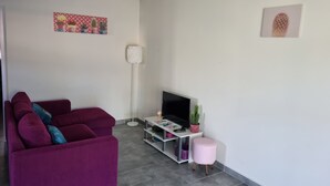 A living room with a purple sofa, a flat-screen TV, a floor lamp, and a small pink stool.