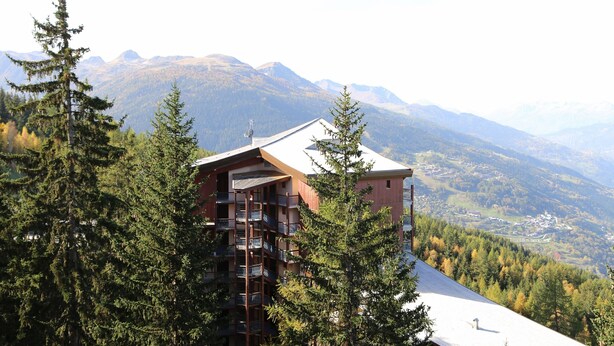 Un edificio de varios pisos con balcones, rodeado de un bosque denso y montañas al fondo.