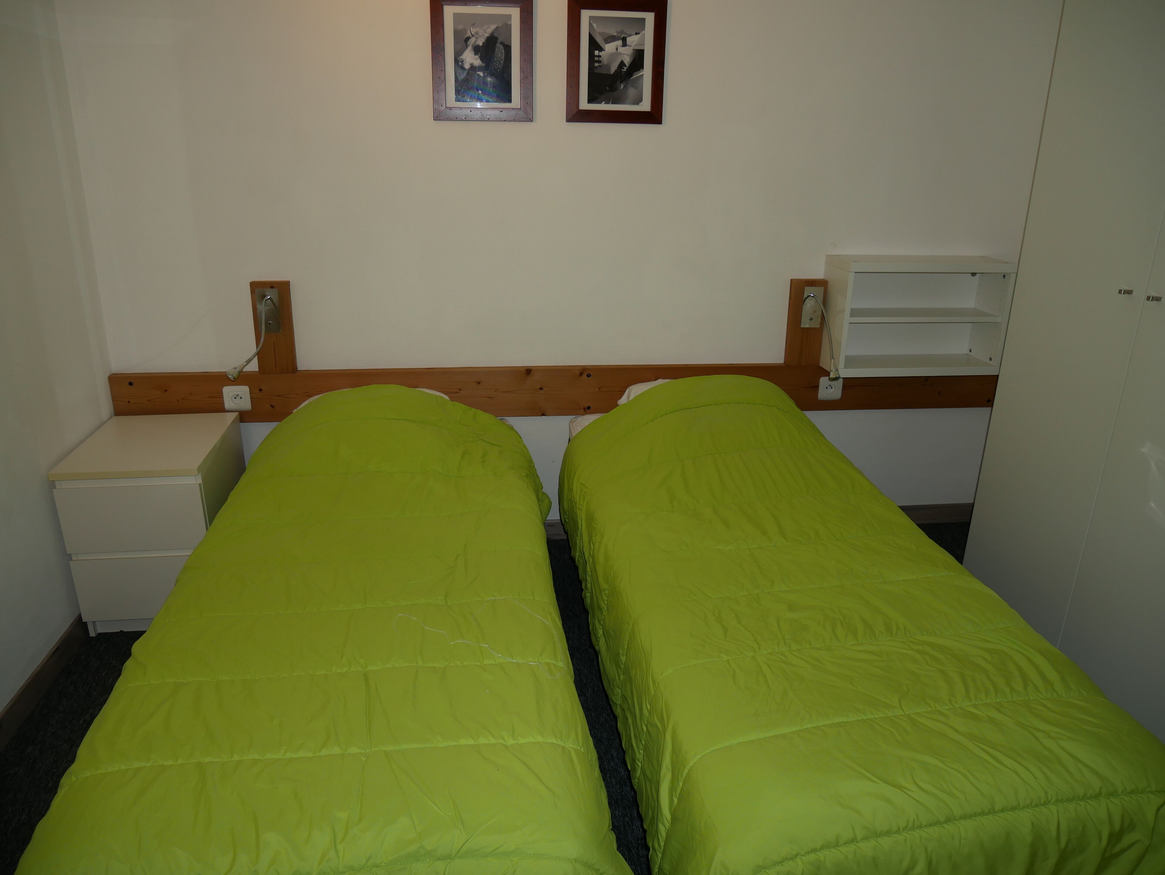 A room with two single beds, a small bedside table, a wardrobe, and two framed pictures on the wall.