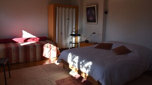 Un dormitorio con una cama grande, una cama más pequeña, una silla, una mesita de noche, una lámpara y un cuadro en la pared.