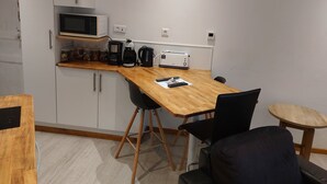Cocina compacta con microondas, tostadora y cafetera sobre la encimera. Hay armarios y una pequeña mesa con sillas.