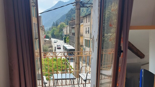 Un balcón con mesa y sillas, vista a una calle con coches y edificios, y una montaña al fondo.