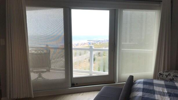 A bedroom with a large sliding glass door leading to a balcony, a bed with a checkered bedspread, and a striped rug on the floor. - Boardwalk Beauty! Two-Story Oceanview Condo (Rehoboth Beach)