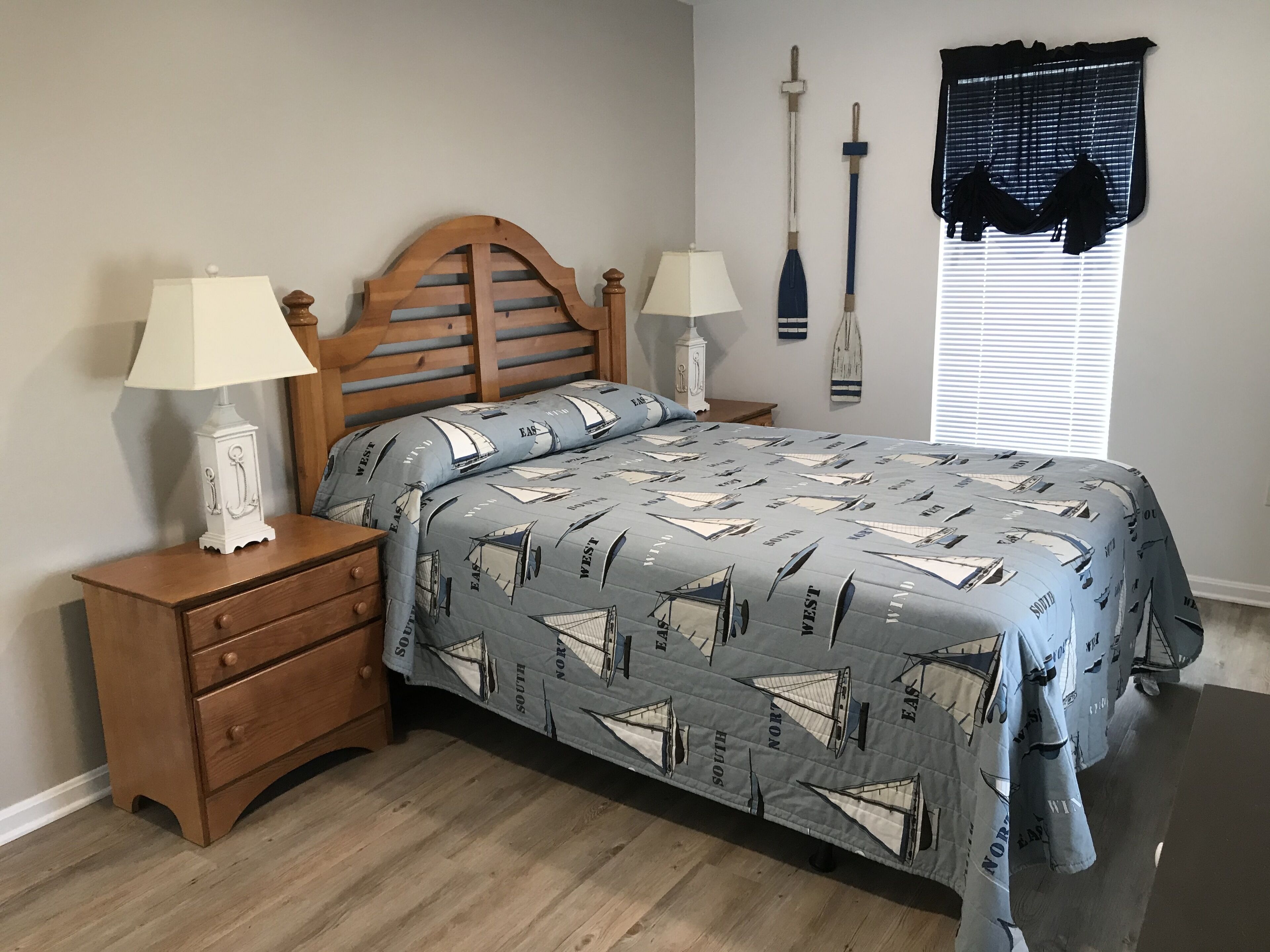 Un dormitorio con cama, mesitas de noche, lámpara, ventana con persianas y un edredón con motivos náuticos.