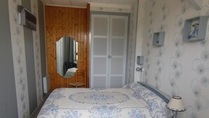 A bedroom with a wooden door, a bed with a floral bedspread, a chair, a bedside table with a lamp, and a wall-mounted shelf with decorative items.