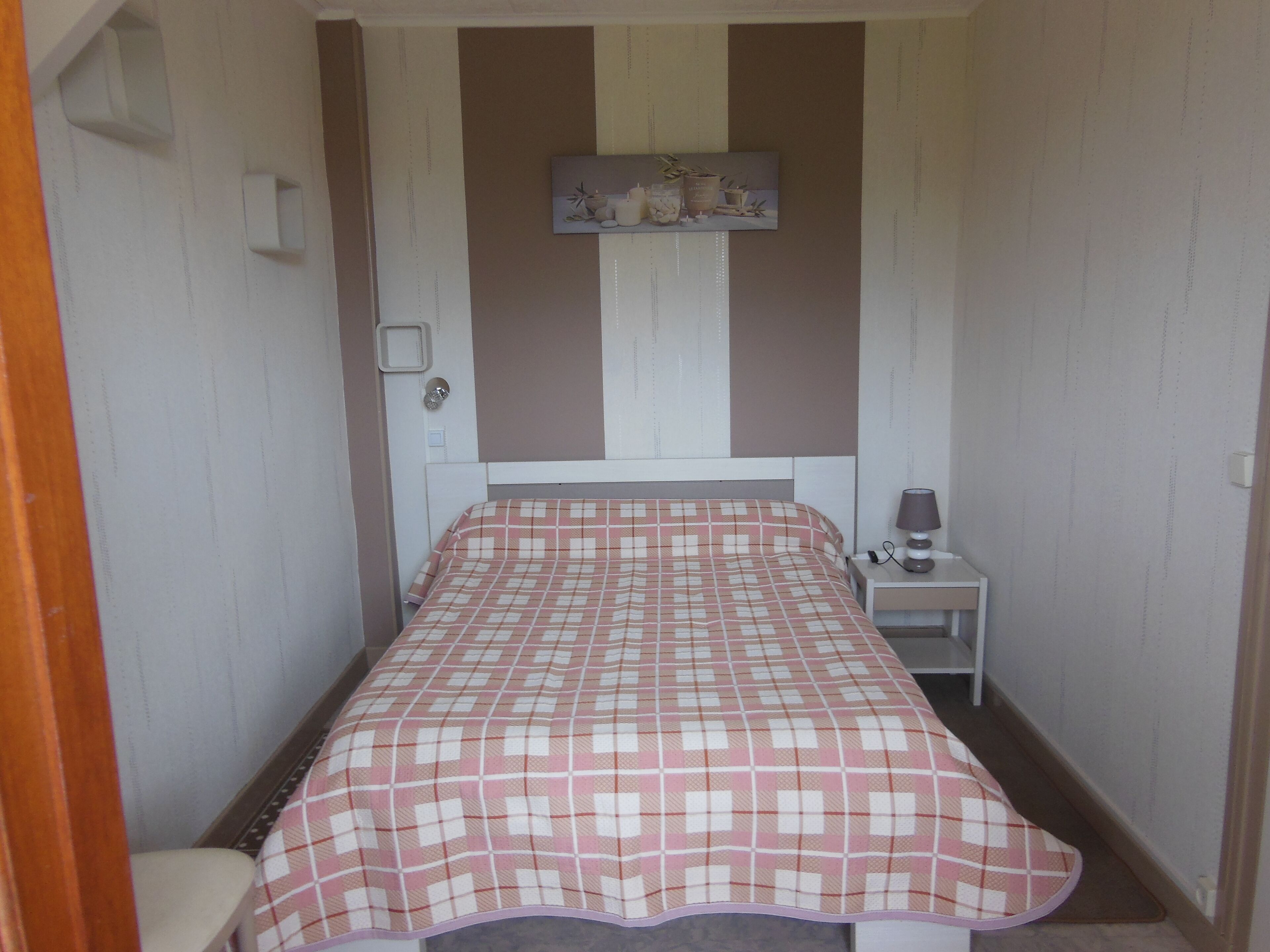 A small, narrow hotel room with a single bed, a bedside table with a lamp, and a wall-mounted shelf with decorative items.
