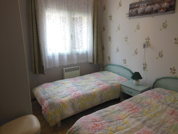 Un dormitorio con dos camas individuales, una ventana con cortinas translúcidas, un aire acondicionado montado en la pared y una mesita con una lámpara.