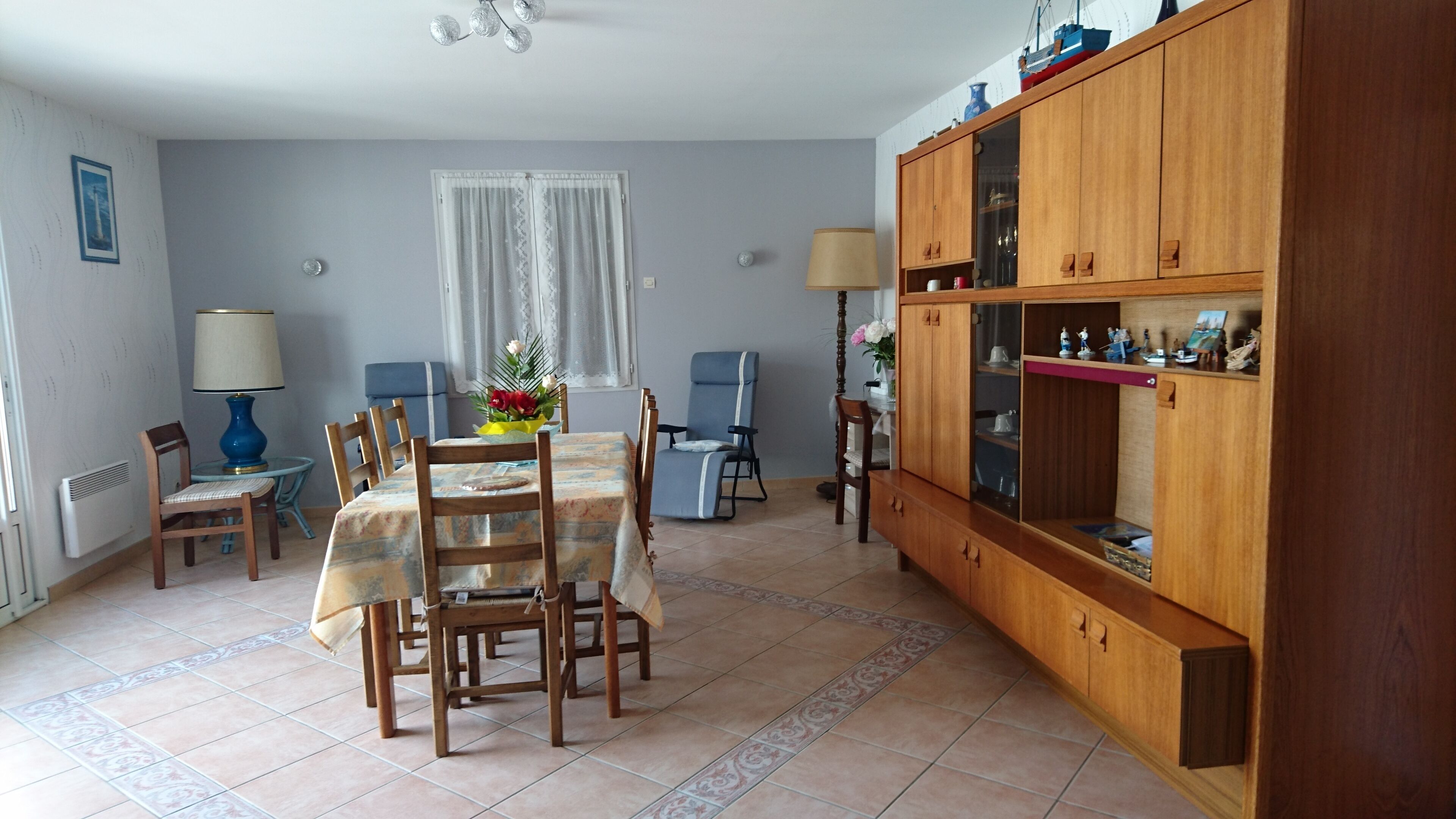 Un comedor con una mesa dispuesta para cuatro personas, rodeada de sillas, un aparador de madera con puertas de vidrio y un ventanal con cortinas.