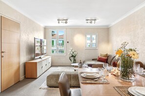Ein Wohnzimmer mit einer Couch, einem Esstisch für vier Personen, einem Fernseher und einem großen Fenster mit Blick auf Gebäude.