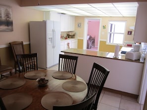 Ein Essbereich mit einem Holztisch, Stühlen und Untertassen. Im Hintergrund ist eine Küche mit einem Kühlschrank zu sehen.