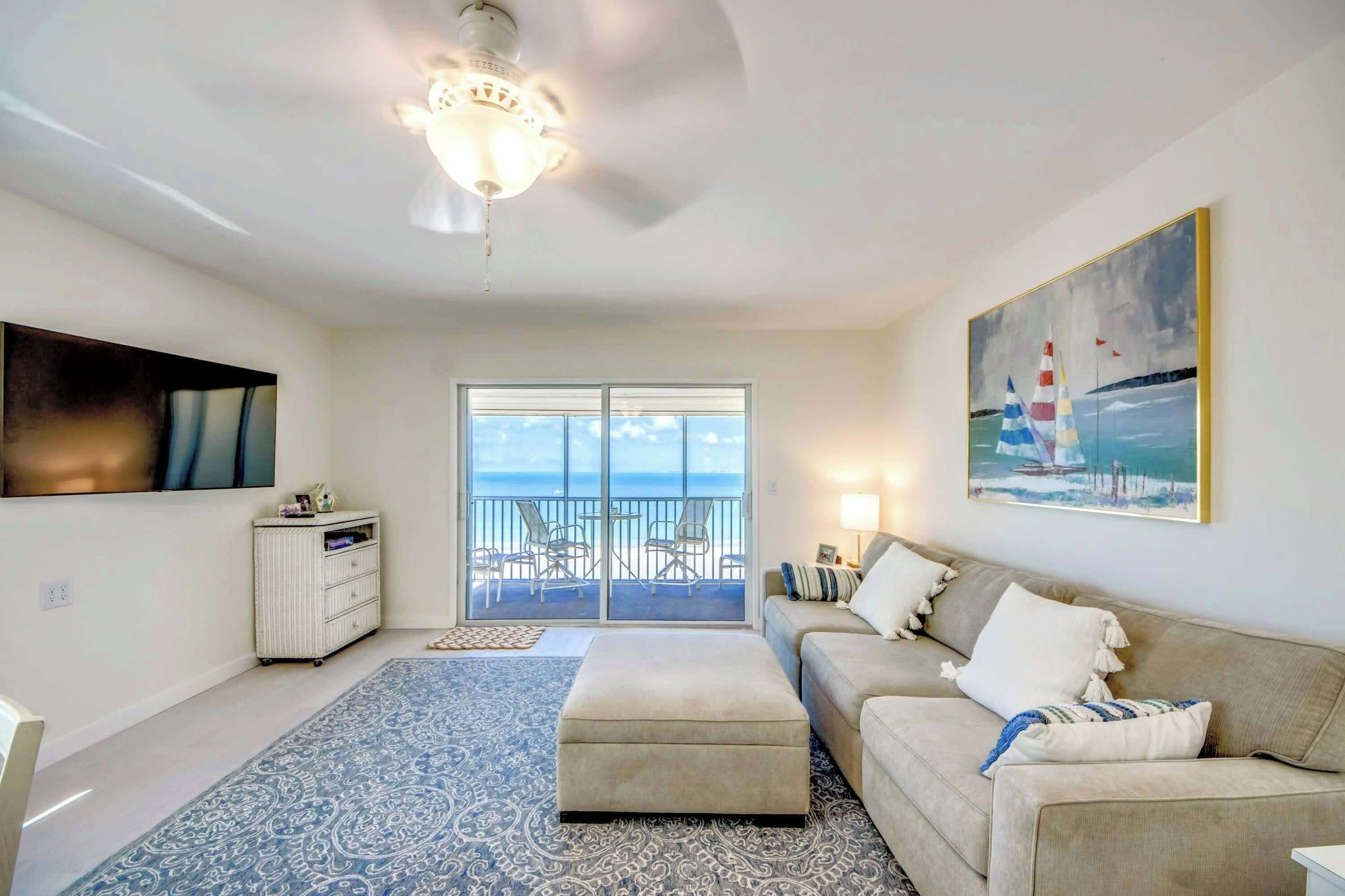 A living room with a sofa, ottoman, and a ceiling fan. There is a large window with a view of the ocean.