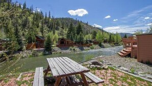 A wooden cabin with a picnic table by a river, surrounded by dense forest. - Cozy Cabin on the River. 1 Bed 1 Bath Patio w/  BBQ (Breckenridge)