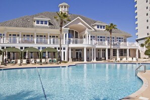 A large, clear swimming pool with lounge chairs and umbrellas, surrounded by a multi-story building with balconies and a tower.