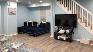 A living room with a dark blue sectional sofa, a glass coffee table, a chandelier, a flat-screen TV, and a staircase.