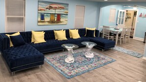 A living room with a dark blue sectional sofa, yellow cushions, a glass coffee table, and a beach-themed painting on the wall.