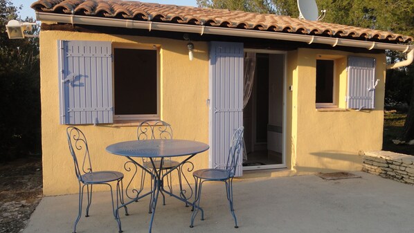 A small building with a tiled roof, two windows with shutters, and a patio with a table and chairs.