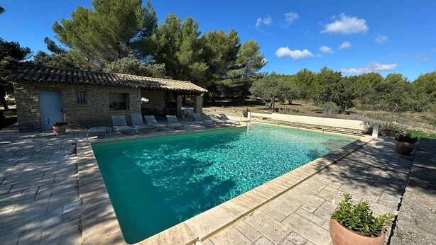 A rectangular swimming pool with clear water, surrounded by stone tiles and a stone building with a tiled roof.