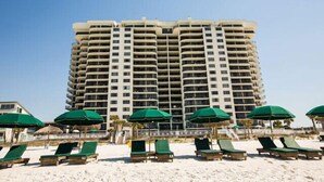 A tall hotel with multiple balconies, a sandy beach in the foreground with green umbrellas and lounge chairs, and a clear blue sky.
