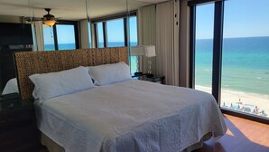 A large bed with a textured headboard, a bedside table with a lamp, and a view of the beach through large windows.