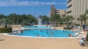 Zona de piscina con varias piscinas, palmeras y un edificio grande al fondo.