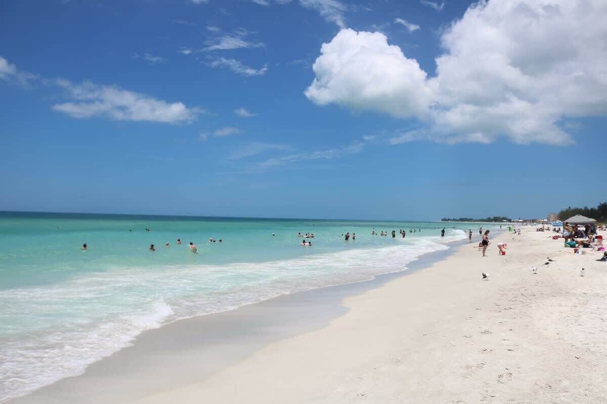 Une plage avec une eau turquoise et limpide, des gens qui se baignent et prennent des bains de soleil, et quelques parasols de plage.