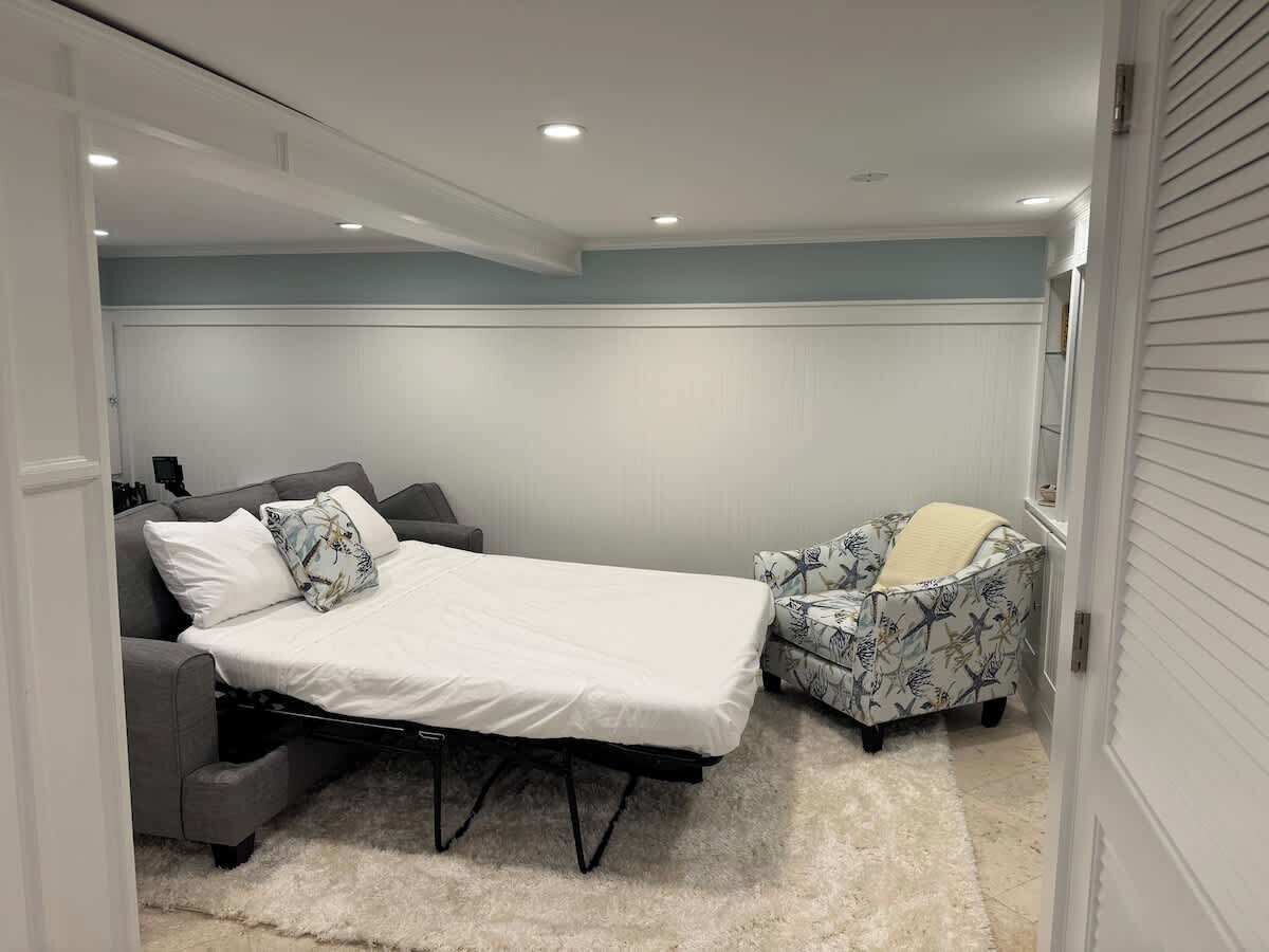 A bedroom with a bed, a grey sofa, a patterned armchair, and a white door.