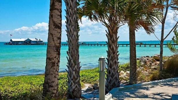 A wooden boardwalk with palm trees, a pier extending into turquoise water, and a building with a flag. - Gulf Front Guest House (Anna Maria)