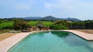 A clear swimming pool with a curved edge, surrounded by a landscaped garden with rocks and plants, set against a backdrop of green fields and distant mountains. - Pleasant, sunny villa (Vaison la Romaine)