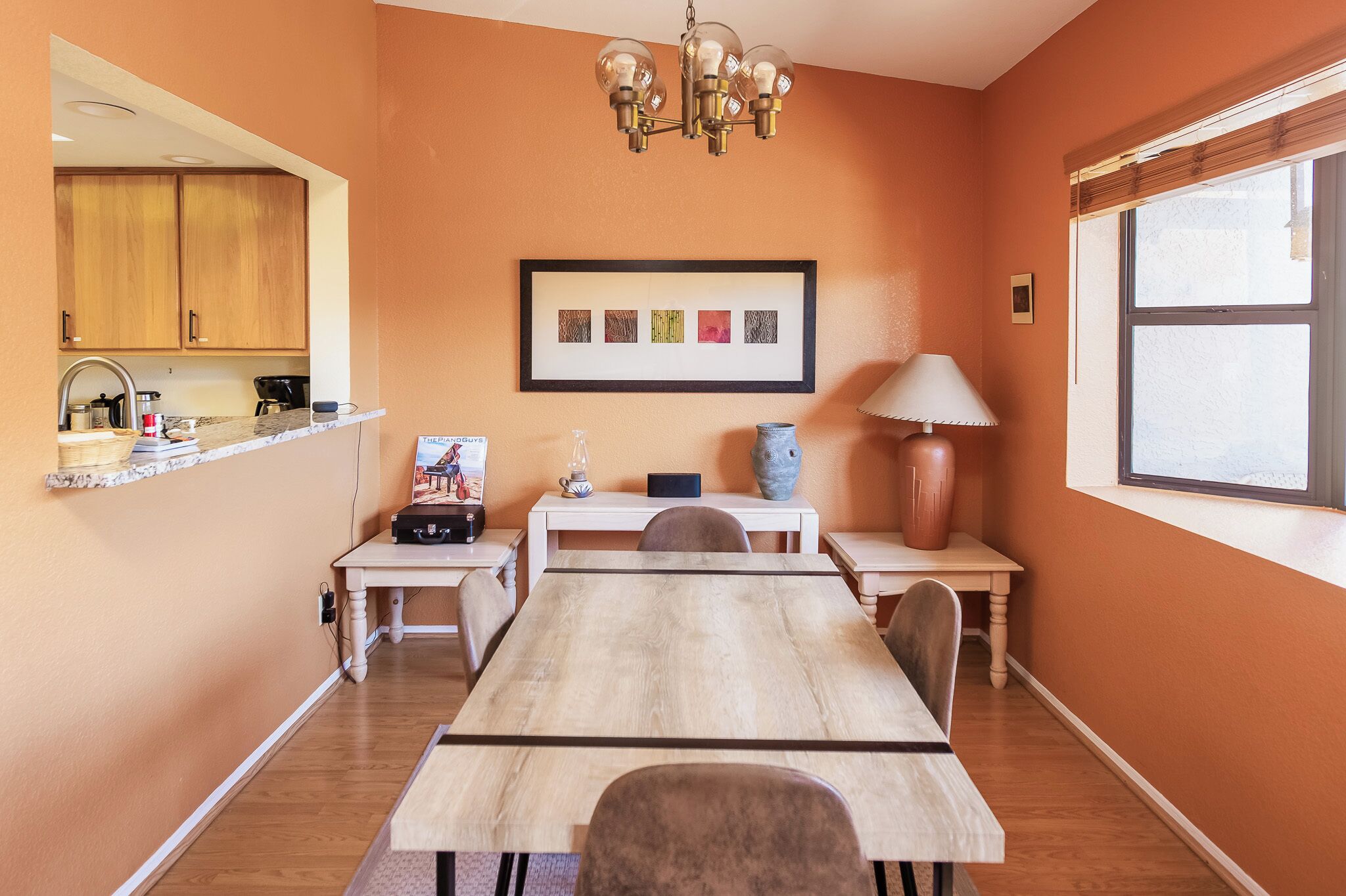 A dining area with a wooden table, chairs, a chandelier, a framed picture, and a window.