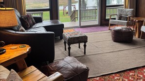 Habitación acogedora con un ventanal grande con vistas a un lago, amueblada con un sofá de cuero, una mesa de madera y otomanas.